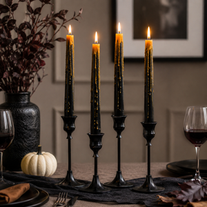 Four black taper candles in matching holders, lit and set on a table runner with autumn decor including a small pumpkin, wine glasses, and a dark vase in the background.