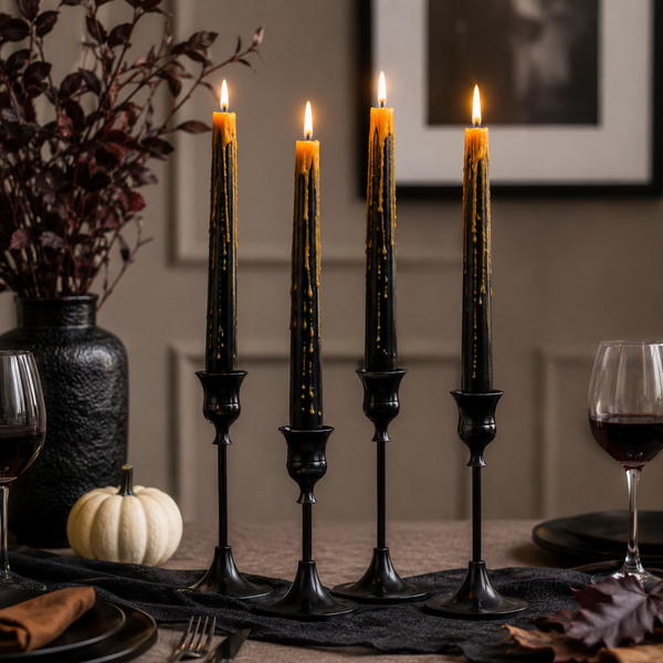 Four black taper candles in matching holders, lit and set on a table runner with autumn decor including a small pumpkin, wine glasses, and a dark vase in the background.