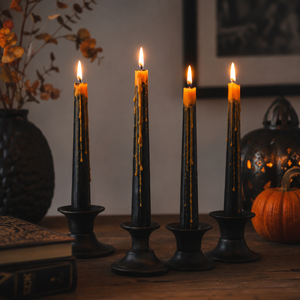 Four black taper candles in matching holders, lit and arranged on a wooden table with a warm orange glow, surrounded by seasonal decor including a pumpkin, books, and a dark vase.