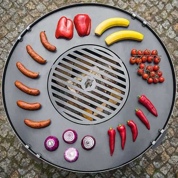 A black steel grill plate with a grate, displayed on a stone surface, surrounded by various vegetables including carrots, tomatoes, and red onions, along with pieces of wood for grilling.