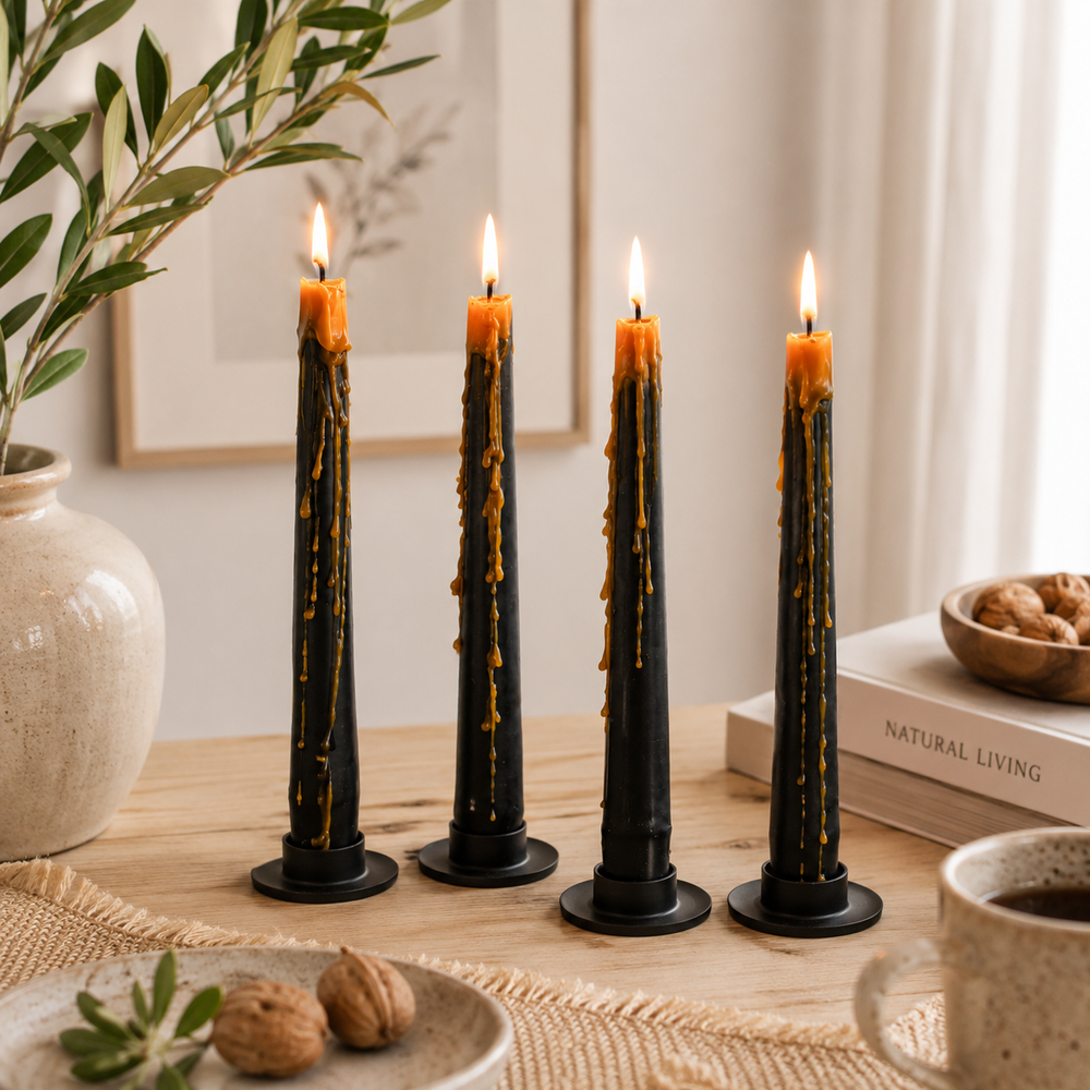 Four black taper candles with melted golden wax drips and lit flames, arranged on a wooden table in a warm, styled interior setting with books, a ceramic vase, and small food props.