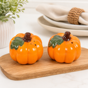 Two orange pumpkin-shaped salt and pepper shakers with green leaves and brown stems, displayed on a wooden cutting board in a kitchen setting.