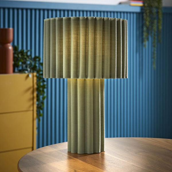 Green pleated lamp on a wooden table with a blue wall and yellow cabinet in the background