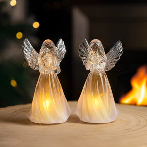 Two angel-shaped lights on a wooden surface with a warm glow, blurred lights and fire in the background.