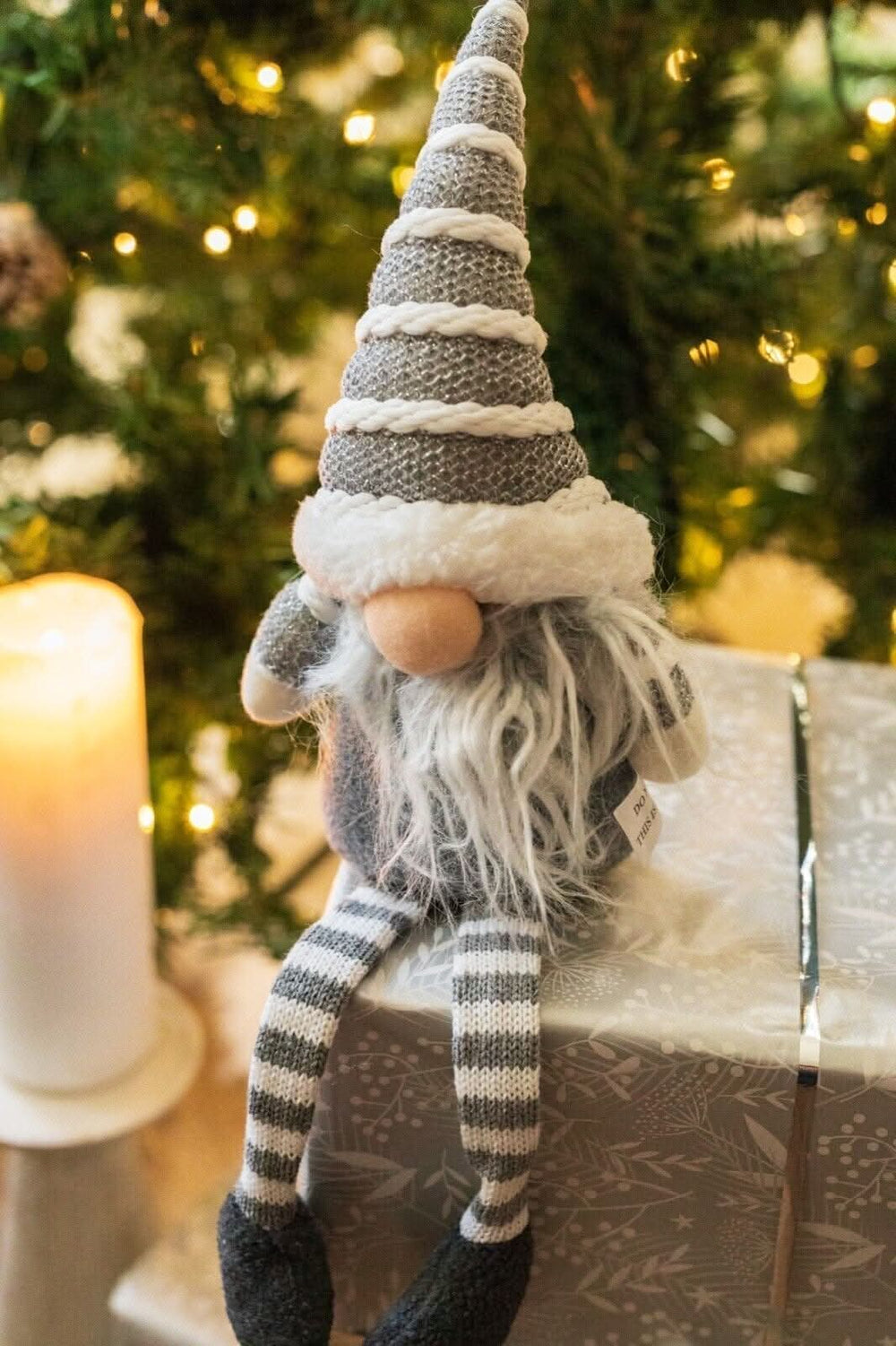 A plush Christmas gnome decoration with a striped hat and gloves, sitting atop a wrapped gift box with a Christmas tree in the background.
