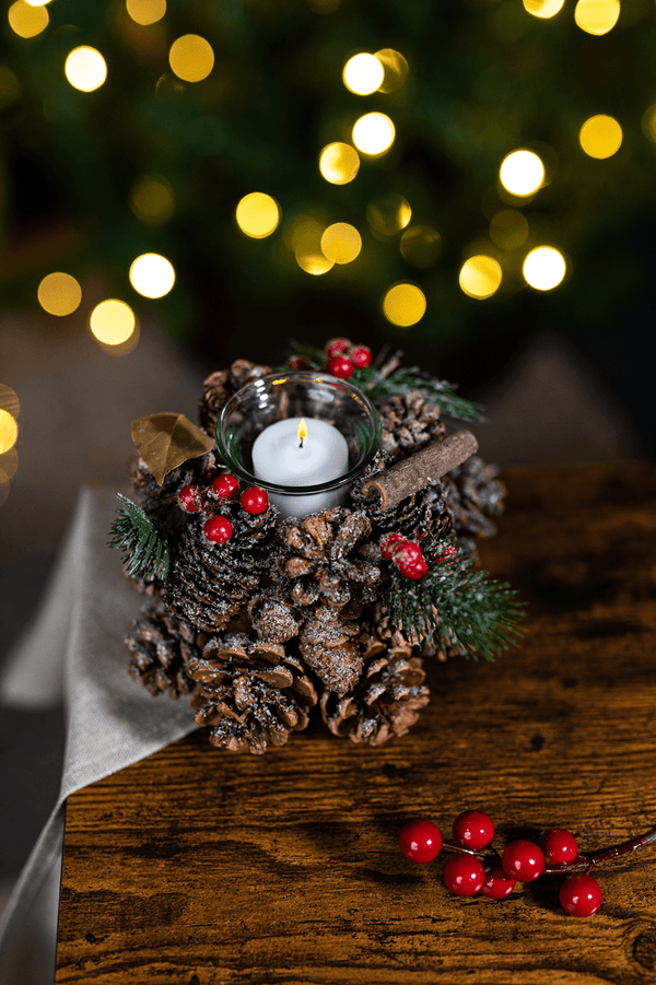 A Christmas-themed tea light candle holder decoration featuring pinecones, red berries, and a white candle, set on a wooden surface with a blurred Christmas tree in the background.