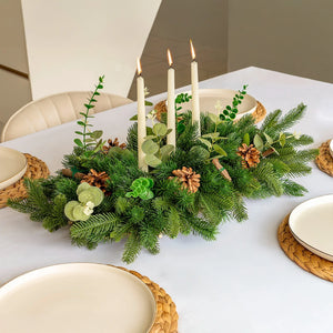 Table setting with a festive centerpiece of greenery, candles, and pinecones on a white tablecloth.