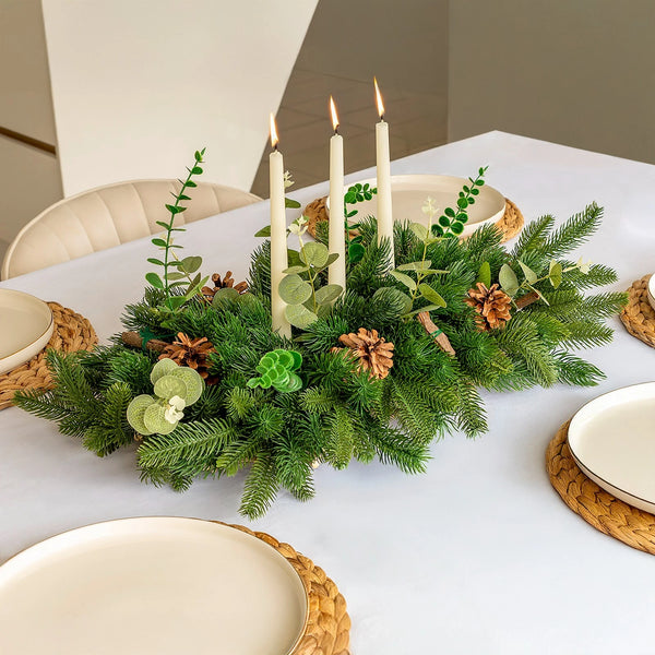 Table setting with a festive centerpiece of greenery, candles, and pinecones on a white tablecloth.