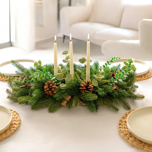 Decorative table setting with greenery, candles, and pinecones on a white surface.