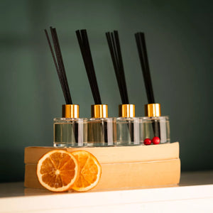 Four diffuser bottles with black sticks on a stack of books with oranges and berries, against a green background.