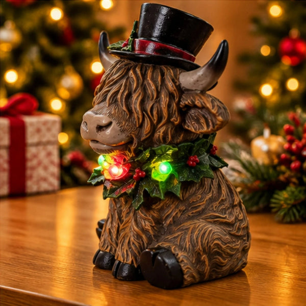 A Highland cow figurine decorated for Christmas, wearing a black top hat with a red band and a green holly garland with red lights, sitting on a wooden table with a blurred festive background.