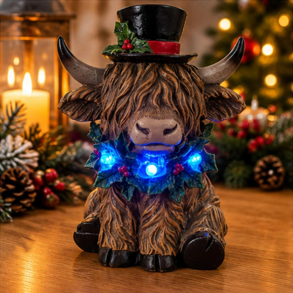 A Highland cow figurine with a black top hat, red band, and a glowing blue LED Christmas holly collar, sitting on a wooden table with a warm, festive background of candles and a blurred Christmas tree.