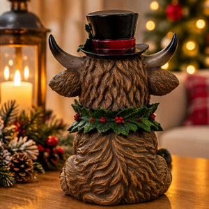A decorative Highland cow figurine with brown textured fur, a black top hat with a red band, and a green holly wreath with red berries, shown on a wooden table in a warm holiday setting with a lantern and blurred Christmas tree in the background.