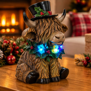 A decorative Highland cow figurine with a lit Christmas wreath and black top hat, sitting on a wooden table in a cozy holiday setting with a blurred fireplace in the background.