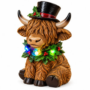 A Highland cow holiday figurine with a black top hat, red band, green holly leaves, and multicolor LED lights around its neck, set against a plain white background.