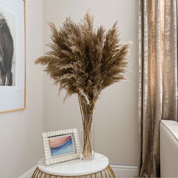 Dried pampas grass in a clear vase on a small round table with a framed picture and curtain in the background.