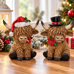 Two Highland cow holiday figurines sitting on a wooden table, one wearing a red Santa hat and candy cane, and the other wearing a black top hat with a red band and holly, with Christmas decorations and a blurred Christmas tree in the background.