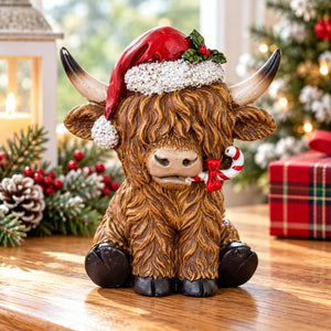 A decorative Highland cow figurine with a brown shaggy coat, black hooves, curved horns, a red Santa hat with white trim, and a candy cane in its mouth, displayed on a wooden table with holiday decorations in the background.