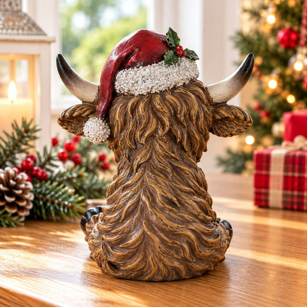 A decorative Highland cow figurine shown from the back, wearing a red Santa hat with white trim and a holly sprig, sitting on a wooden table with a blurred Christmas tree and holiday decorations in the background.