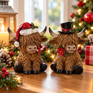 Two Highland cow holiday figurines sitting on a wooden table, one wearing a red Santa hat and candy cane, and the other wearing a black top hat with a red band and holly