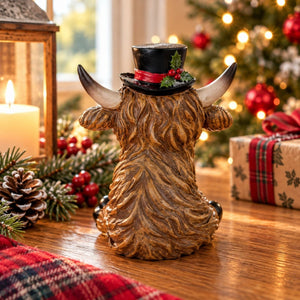 A decorative Highland cow figurine shown from the back, wearing black hat with a red band and a holly, sitting on a wooden table with a blurred Christmas tree and holiday decorations in the background.