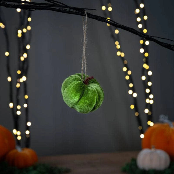 Green felt apple ornament hanging on a branch with blurred lights and pumpkins in the background