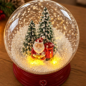 A lit Christmas snow globe with a red base, featuring miniature evergreen trees, a Santa Claus figure, and a red mailbox surrounded by glowing warm lights on a wooden surface.