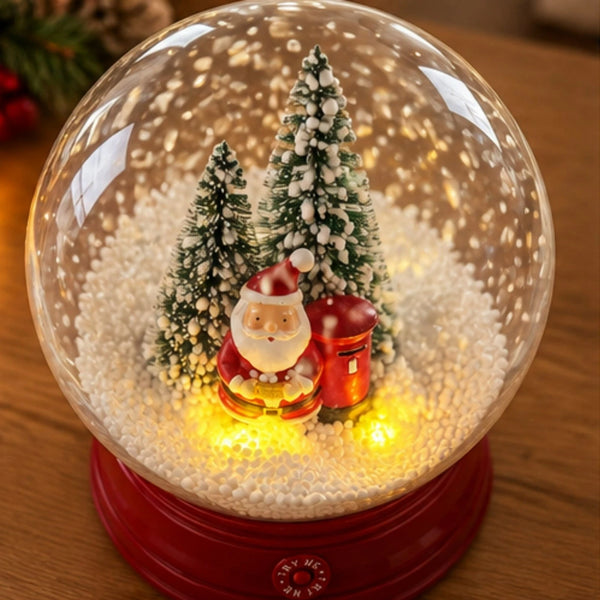 A lit Christmas snow globe with a red base, featuring miniature evergreen trees, a Santa Claus figure, and a red mailbox surrounded by glowing warm lights on a wooden surface.