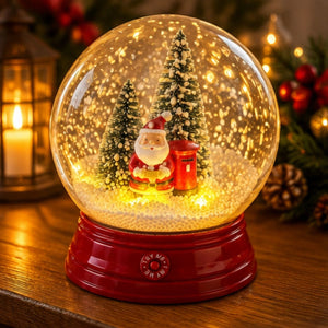 A festive Christmas snow globe with a red base, glowing warm lights inside, a Santa Claus figure, and small decorated trees, displayed on a wooden surface with holiday decor in the background.