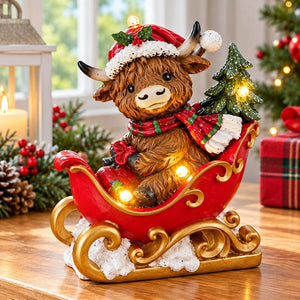 A festive Highland cow figurine sitting in a red sleigh with gold trim, surrounded by holiday lights, a small Christmas tree, and white faux snow, displayed on a wooden surface with a cozy Christmas background.