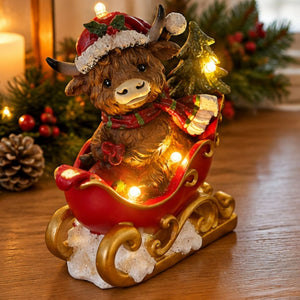 A festive Christmas bull figurine sitting in a red sleigh with gold trim, decorated with warm white string lights, a small lit Christmas tree, and a Santa hat, displayed on a wooden surface with holiday greenery in the background.