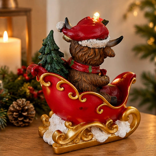 A festive Christmas figurine of a brown dog in a red Santa hat sitting in a red sleigh with gold trim, a small green tree, and white faux snow, displayed on a wooden surface with warm holiday decor in the background.