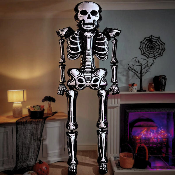 A large black and white skeleton-shaped floor decoration stands in a decorated living room with a lit fireplace, spiderweb wall decor, and Halloween-themed props.