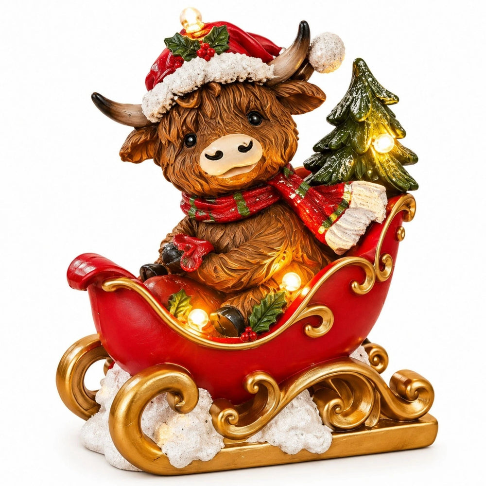 A festive Highland cow figurine sitting in a red sleigh with gold trim, surrounded by holiday lights, a small Christmas tree, and white faux snow, displayed on a wooden surface with a cozy Christmas background.