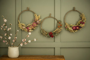 Three decorative wreaths on a green wall with a vase of flowers on a wooden surface.