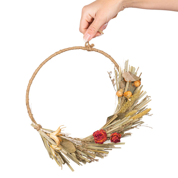 Hand holding a decorative wreath with dried elements and red flowers on a white background