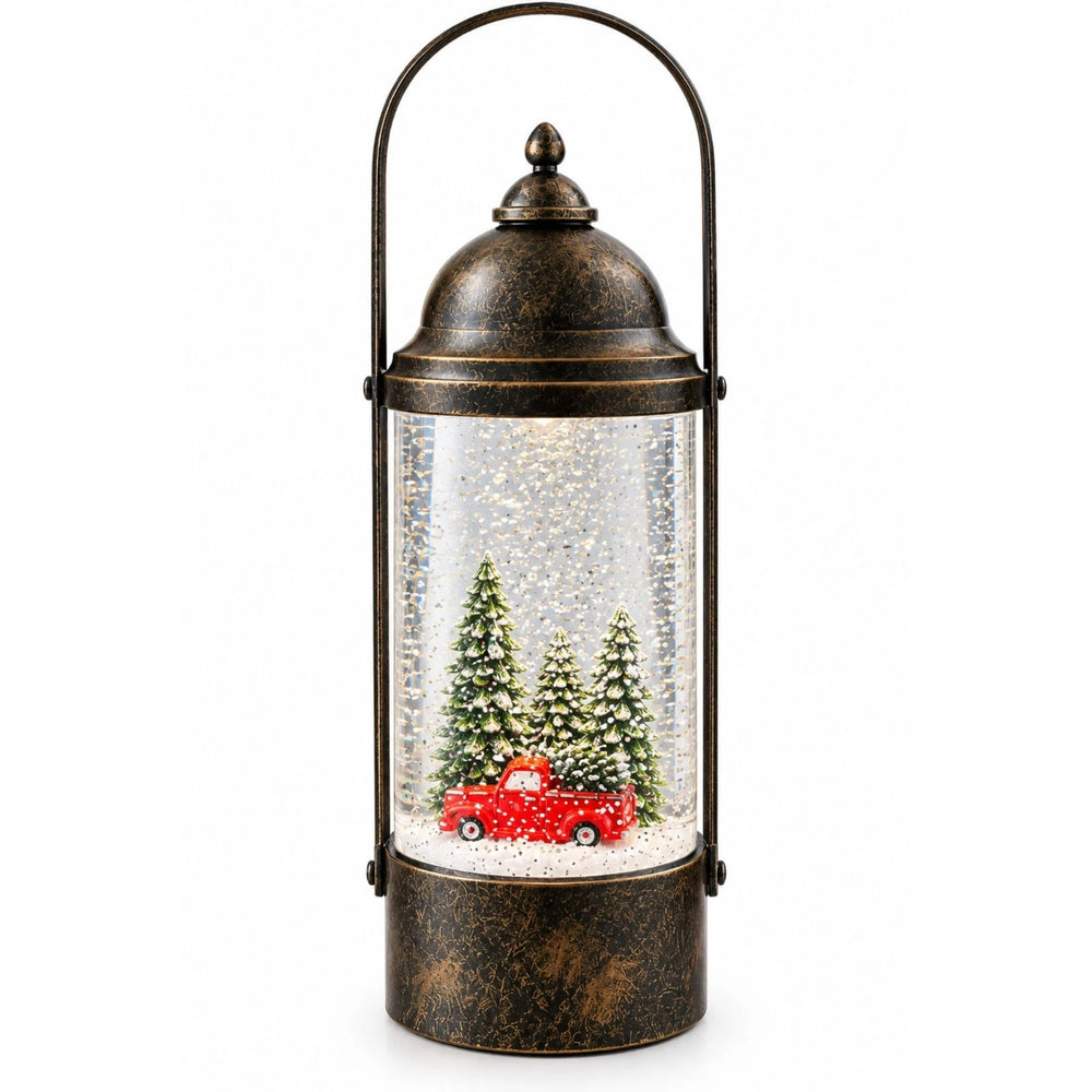 A rustic metal lantern-style holiday table lamp sits on a wooden table in a cozy living room. The lamp has a distressed bronze finish, a clear glass body filled with white glitter and miniature Christmas trees, and a small red pickup truck near the bottom.