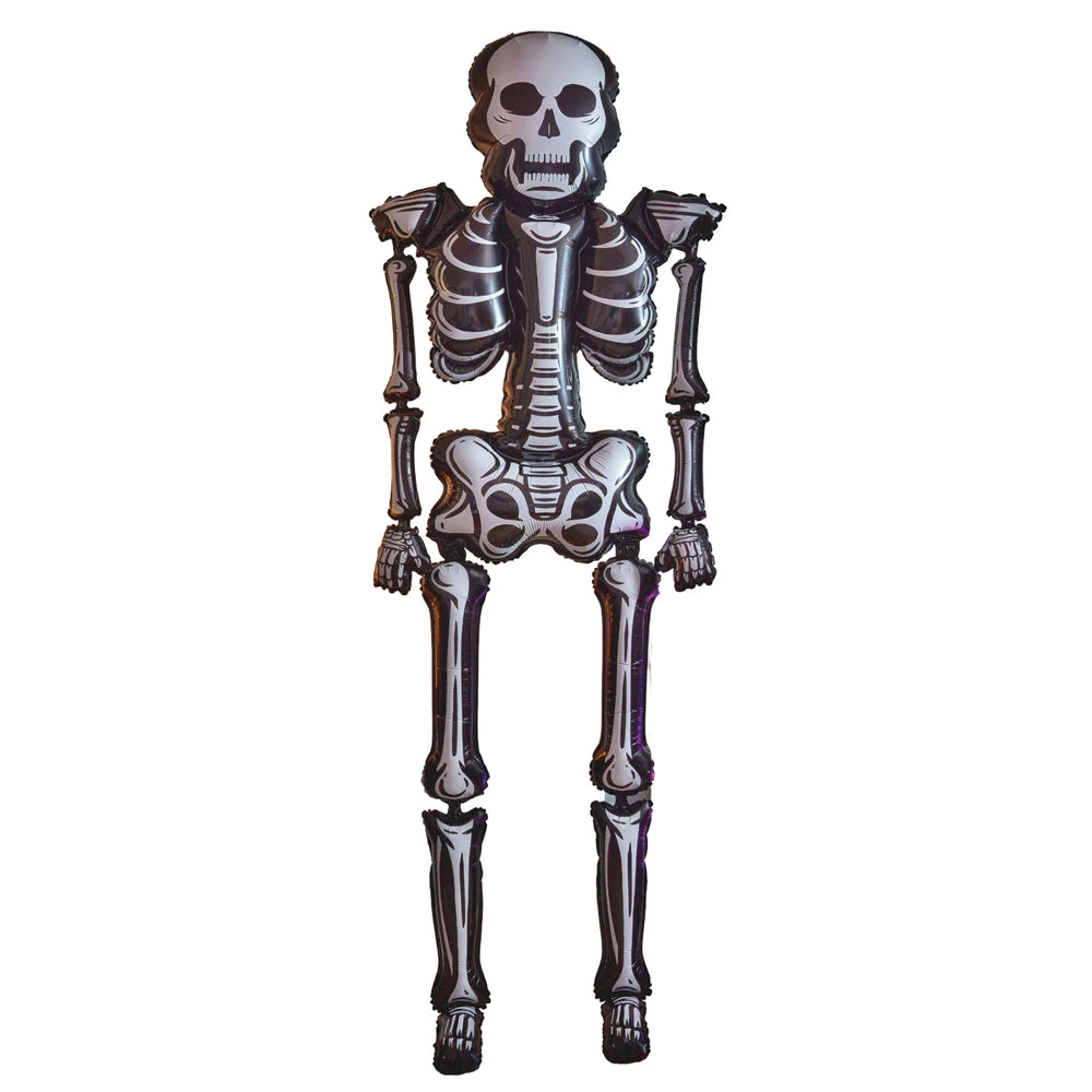 A large black and white skeleton floor decoration stands in a cozy, Halloween-decorated living room with a fireplace, pumpkins, and themed pillows.