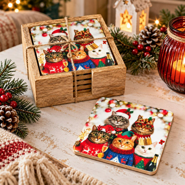 A set of square coasters with a festive Christmas cat illustration, shown with one coaster standing in a wooden box and another lying flat on a rustic table. The scene is decorated with pine branches, red berries, a lit candle in a glass jar, and a knitted blanket.