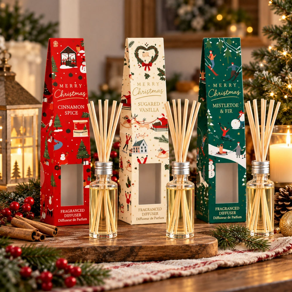 Three Christmas-themed reed diffusers with their decorative packaging displayed on a wooden tray, surrounded by holiday decorations including candles, pine branches, cinnamon sticks, and ornaments.