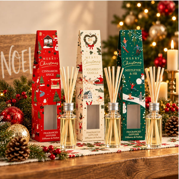 Four Christmas-themed reed diffuser bottles with wooden reeds are arranged on a wooden table in front of their decorative packaging boxes. The products include Cinnamon Spice, Sandalwood Vanilla, and Mistletoe & Fir scents.