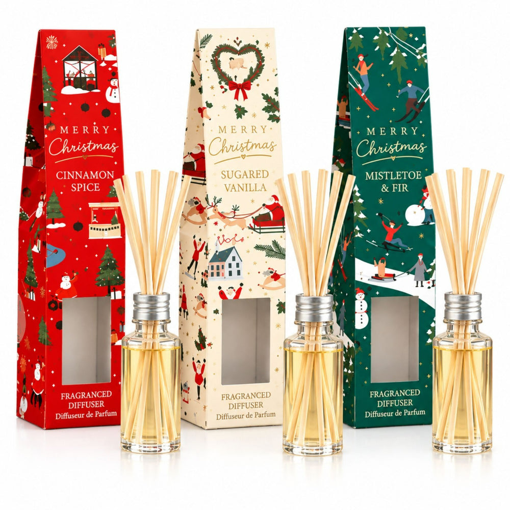 Three Christmas-themed reed diffusers with their decorative packaging displayed on a wooden tray, surrounded by holiday decorations including candles, pine branches, cinnamon sticks, and ornaments.