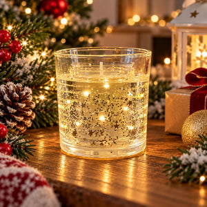 A clear glass candle holder filled with warm golden liquid and embedded fairy lights, styled on a wooden table with Christmas decorations including pine branches, a pinecone, red berries, a lantern, and a gift box in the background.