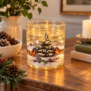 A clear glass candle jar with a warm white LED light inside, featuring a printed winter village scene with a Christmas tree, cabins, snow, and falling leaves