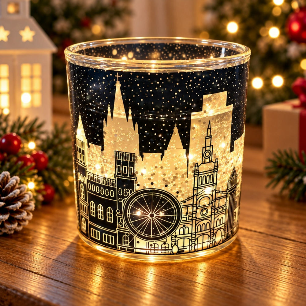 A clear glass candle holder with a black and gold Christmas cityscape design, glowing with warm light on a wooden table surrounded by holiday decorations.