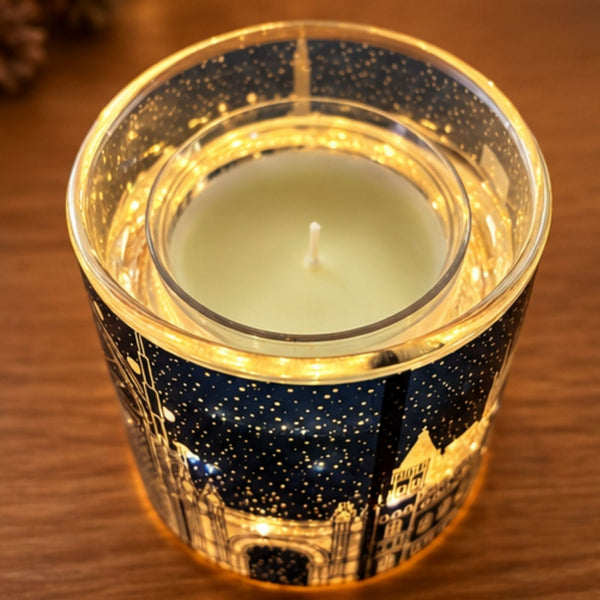 A lit tealight candle in a clear glass holder with a navy blue and gold cityscape design, photographed on a wooden surface.