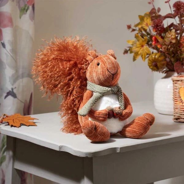 Plush squirrel toy with a scarf on a white surface, surrounded by autumn decorations.