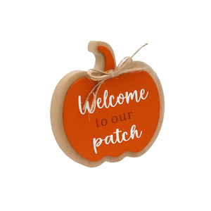 A wooden pumpkin-shaped decorative sign with an orange face, natural wood border, white script text reading 'Welcome to our patch,' and a small jute twine bow on a stem, shown on a plain white background.