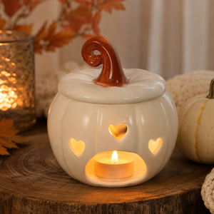A white ceramic pumpkin-shaped candle holder with a brown stem, featuring heart-shaped and window cutouts that glow warm yellow from a tea light candle inside. It sits on a rustic wooden slice with autumn decor in the background.