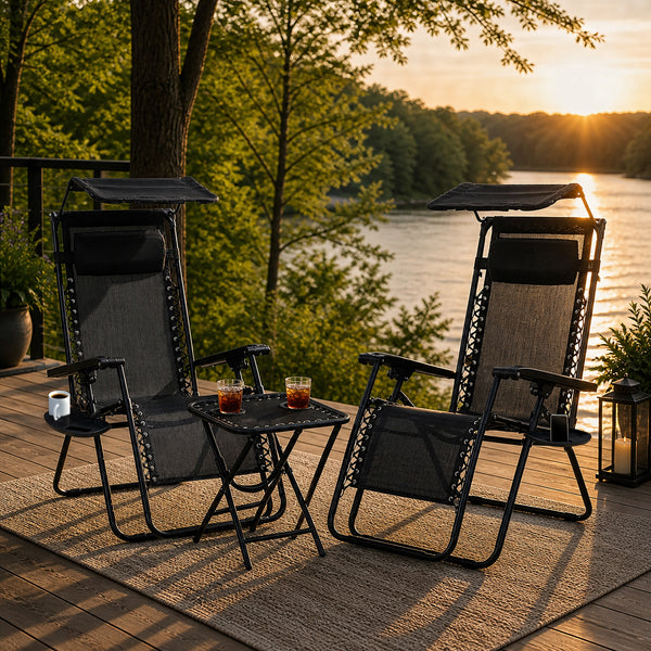 Two black patio chairs with a small table on a wooden deck overlooking a river at sunset.
